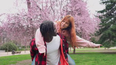 Neşeli ırklar arası çift neşeli sırtımda gezintiyi canlı bahar günü çiçek açan park ağaçlarının arasında genç aşklarını ve tasasız ilişkilerini sergiliyorlar.