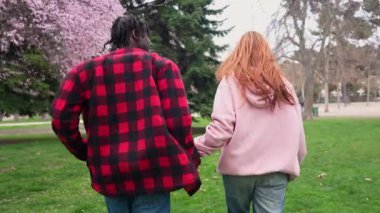 Irklar arası genç çift birlikte koşarken samimi bir anı paylaşıyorlar, yeşil park manzarasında el ele tutuşuyorlar, kaygısız romantik karşılaşmada neşe ve bağlantıyı ifade ediyorlar.