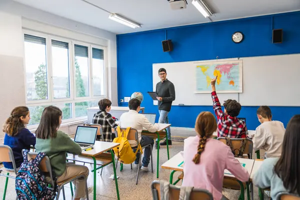 Coğrafya öğretmeni genç öğrencilere ders veriyor, bir öğrenci modern sınıfta bir soruyu cevaplamak için elini kaldırıyor.