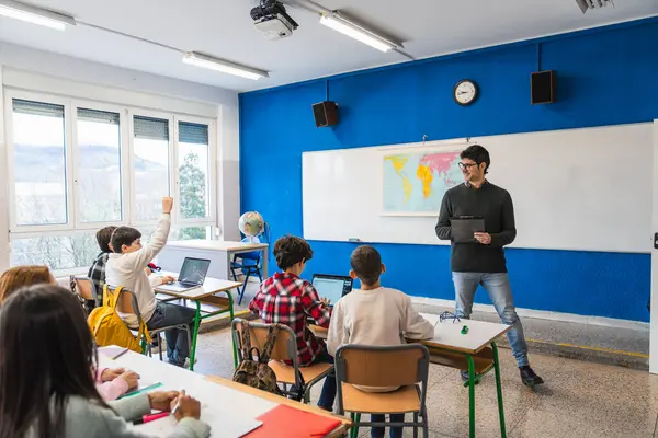 İlkokul öğrencileri, modern bir sınıftaki öğretmenin sorularını cevaplamak için dizüstü bilgisayarlarıyla masalarında oturuyor ve ellerini kaldırıyorlar.