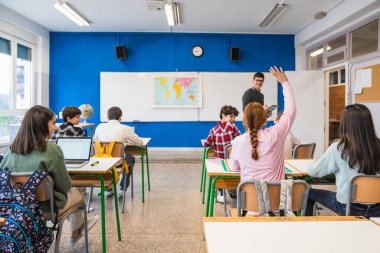 İlkokul öğrencileri öğretmenle coğrafya öğreniyor ve modern sınıfta soruları cevaplamak için el kaldırıyorlar.