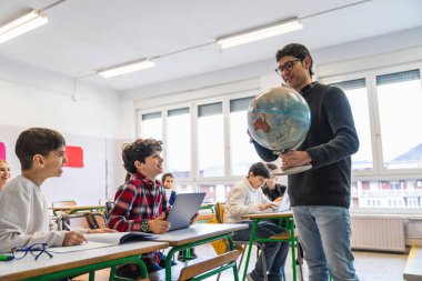 Coğrafya öğretmeni coğrafya dersi sırasında ilkokul öğrencilerine bir küre gösteriyor.