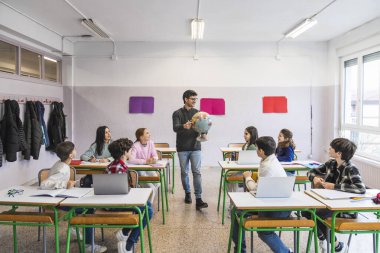 Coğrafya öğretmeni, dünya kültürleri ile ilgili interaktif bir sınıf dersi sırasında bir küre sunarak ilkokul öğrencilerini meşgul ediyor.