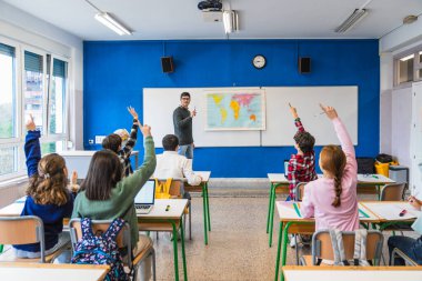 Bir grup ilkokul öğrencisi coğrafya dersinde öğretmenin sorusuna cevap vermek için el kaldırıyor ve dünya haritasını öğreniyorlar.