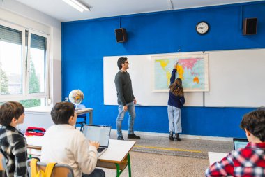 Coğrafya öğretmeni modern bir sınıfın duvarındaki dünya haritasını gösteren öğrenciye yardım ediyor.