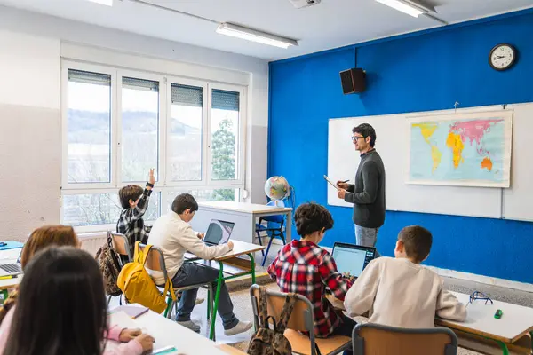 Modern sınıfta dizüstü bilgisayar kullanan ilkokul öğrencilerine not panosu tutan ve coğrafya dersi veren öğretmen.