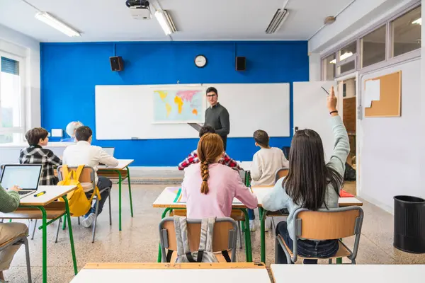 Bir grup ilkokul öğrencisi coğrafya dersinde el kaldırırken, öğretmen dersi anlatıyor.