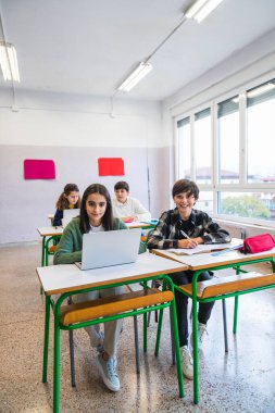 İlkokul öğrencileri, modern sınıflarda dizüstü bilgisayar kullanarak ve notlar alarak sıralarında oturuyorlar.