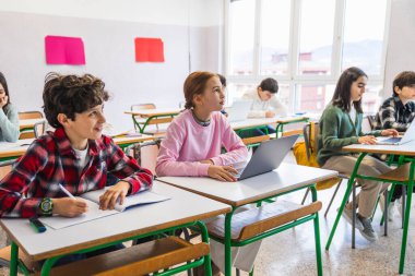 Çeşitli ilkokul öğrencileri sıralarında oturup dizüstü bilgisayar ve not defteri kullanarak öğretmeni dikkatle dinliyorlar.