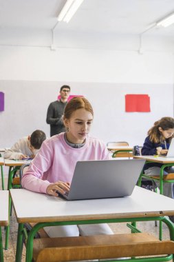 Sınıfta dizüstü bilgisayar kullanan odaklanmış bir öğrenci, sınav sırasında öğrencileri izleyen bir öğretmen.