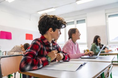 Bir grup ilkokul öğrencisi sıralarında oturuyor, öğretmenlerini dinliyor ve ders sırasında yeni kavramlar öğreniyorlar.