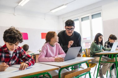 Ders sırasında dizüstü bilgisayar kullanan kız öğrenciye asistanlık yapan öğretmen ve diğer öğrenciler arka planda çalışıyor.