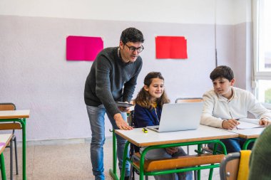 Ders sırasında sınıfta dizüstü bilgisayar kullanan ilkokul öğrencilerine yardımcı olan öğretmen.