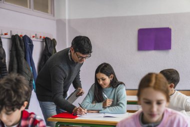 Öğretmen bir kız öğrenciye sınıfta egzersiz yaptırırken, diğer öğrenciler de arka planda öğreniyor.