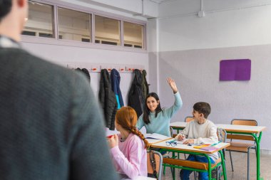 Öğrenci kız, sınıfta ders sırasında öğretmeninin sorusuna cevap vermek için elini kaldırıyor. Katılma ve öğrenme hevesini gösteriyor.