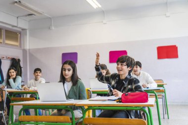 Oğlan sınıfta el kaldırırken, kız bilgisayar kullanıyor, etkileşimli öğrenme ve modern eğitime katılım sergiliyor