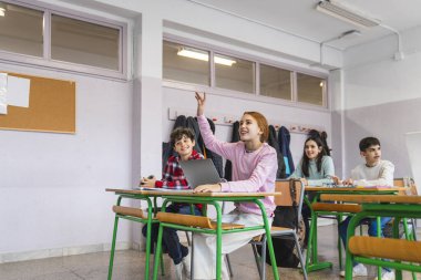 Genç bir kız ders sırasında sınıf arkadaşlarıyla aktif olarak derse katılırken bir soruyu cevaplamak için elini kaldırıyor.
