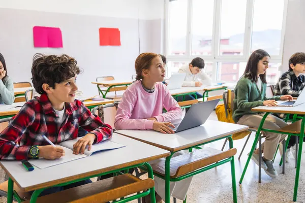 Çeşitli ilkokul öğrencileri sıralarında oturup dizüstü bilgisayar ve not defteri kullanarak öğretmeni dikkatle dinliyorlar.