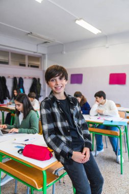 Sınıf arkadaşları derslerine konsantre olurken neşeli bir öğrenci sınıftaki bir masaya yaslanıyor.