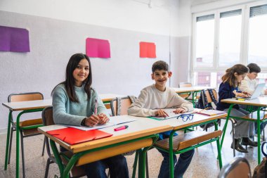 İki genç öğrenci, öğrenme sevincine dalmış, hareketli bir sınıfta, sıralarında çalışmalarına devam ediyor ve gülümsüyor.