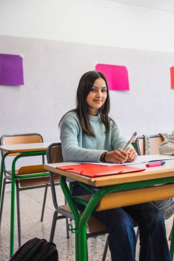 Sınıfta masasında oturan genç bir kız öğrenci, arkasında renkli kağıtlar olan bir not defteri yazıyor.