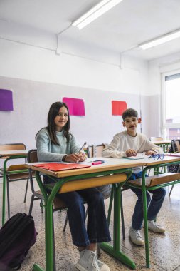 İki ortaokul öğrencisi sınıftaki masada oturup not defterlerine yazı yazıyorlar.