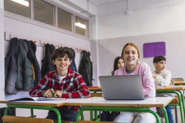 İlkokul öğrencileri sınıfta gülümsüyor ve öğreniyor, kız dizüstü bilgisayar kullanıyor ve oğlan da kitapla çalışıyor.