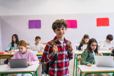 Sınıfta dizüstü bilgisayarlarla çalışan sırt çantası takan gülümseyen bir erkek öğrencinin portresi.