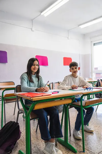 İki ortaokul öğrencisi sınıftaki masada oturup not defterlerine yazı yazıyorlar.