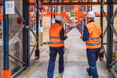 Güvenlik yelekleri ve kask giyen iki depo işçisi büyük bir dağıtım merkezinde envanter ve lojistik işlerini yürütüyorlar.