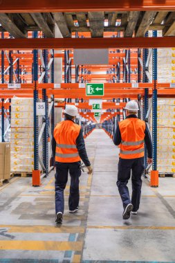 Güvenlik yelekleri ve başlık takan iki depo çalışanı bir lojistik merkezinden geçerek, malları ve operasyonları denetliyor.