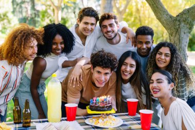Bir grup neşeli çoklu etnik arkadaş açık hava partisinde doğum gününü pasta ve içkiyle kutluyorlar.