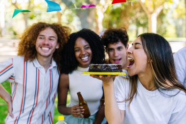 Mutlu arkadaşlar parktaki doğum günü partisini kutlarken genç bir kadın çikolatalı pasta yemek üzere.