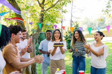 Doğum günü pastası ve renkli parti bayraklarıyla bir parkta alkışlayan ve doğum gününü kutlayan neşeli çoklu etnik arkadaşlar grubu.