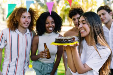 Çikolatalı doğum günü pastası tutan genç bir kadın. Arkadaşlarıyla bir partide gülümsüyor. Parkta renkli flamalarla kutlama yapıyor.