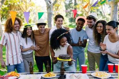 Mutlu çoklu etnik arkadaşlar bir doğum gününü çikolatalı pasta, içki ve atıştırmalıklarla bir parkta süslü piknik masasında kutluyor.