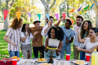 Bir grup neşeli çoklu etnik arkadaş parkta bir doğum günü partisini kutluyor, şarkı söylüyor ve ellerini çırpıyorlar.