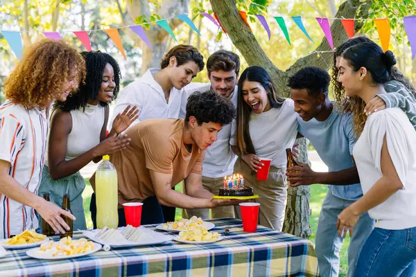 Bir grup neşeli çoklu etnik arkadaş parkta bir piknik masasında pasta ve içkiyle doğum gününü kutluyorlar.