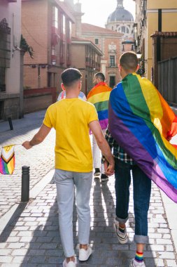 Gün batımında güneşli bir caddede el ele tutuşan, canlı LGBTQ aksesuarlarıyla süslenmiş iki neşeli eşcinsel çiftin arka planı..