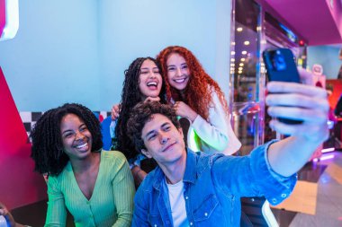 Bir grup mutlu genç arkadaş, renkli bir bowling salonunda ve atari salonunda eğlenirken akıllı telefonlarıyla selfie çekiyorlar.