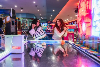 İki mutlu arkadaş, canlı bir oyun odasında hava hokeyi oynayarak arkadaşlıklarını ve oyunun heyecanını kutluyorlar.