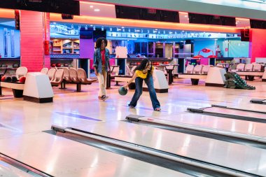 İki genç kadın bovling oynamaktan zevk alıyor, biri topu atıyor ve diğeri renkli bir bowling salonunda izliyor.