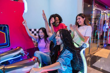 Renkli ve modern bir eğlence merkezinde dört genç kadın birlikte eğleniyor, bir araba yarışı simülatöründe oynuyor ve yarışıyor.