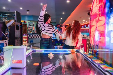 Hava hokeyi oynayan ve zaferlerini renkli bir eğlence salonunda oyun makineleriyle kutlayan iki genç kadın.