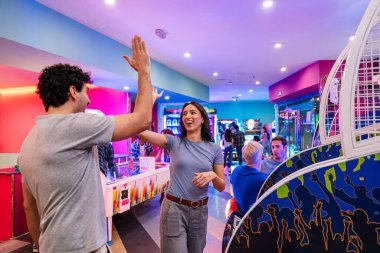 İki arkadaş zaferlerini kutlarken atari oyunlarıyla renkli bir eğlence merkezinde oynuyorlar.