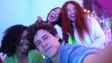 Bir grup çok ırklı arkadaş renkli, neon ışıklı bir odada selfie çekiyorlar ve eğleniyorlar.
