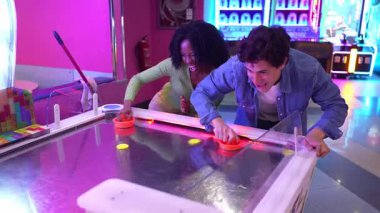 Enerjik çok ırklı çift hava hokeyi oynarken yarışıyor ve gülüyor, renkli oyun ortamında eğlenceli anlar paylaşıyorlar.