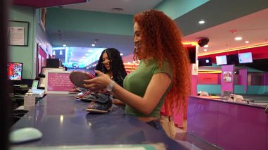 Kadınlar bovling ayakkabılarını tezgahta seçiyor, eğlence için hazırlanırken yavaş çekimde neşeli bir heyecanla hareket ediyorlar.