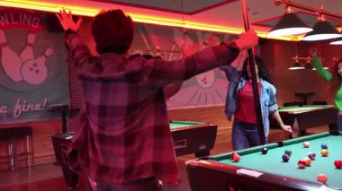 Genç arkadaşlar havuz şutunu kazandıktan sonra çılgınca tezahürat yapıyorlar, zaferi loş ışıklı bovling salonu veya bar ortamında ağır çekim heyecanıyla kutluyorlar.