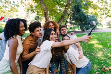 Neşeli genç öğrenciler parkta güneşli bir pikniğin keyfini çıkarırken, bir yandan da aksiyon kamerasıyla selfie çekiyorlar.
